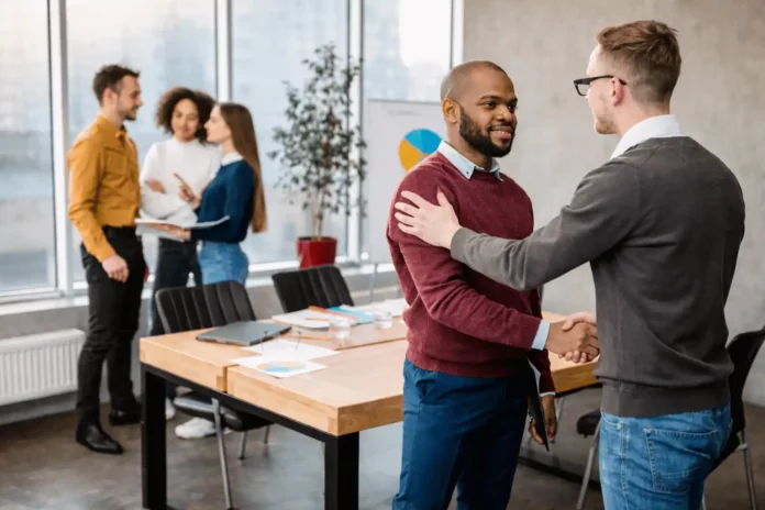 Deux collègues se serrent la main dans un bureau moderne lors d’une intégration dans un nouvel emploi, tandis que d’autres membres de l’équipe discutent en arrière-plan.